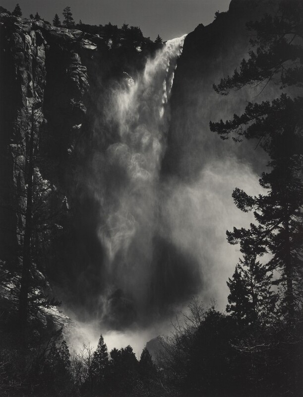 A misty waterfall drops vertiginously from a towering, sheer cliff face that nearly reaches the top edge of this vertical black and white photograph. Spray drifts in hazy clouds down the height and especially around the base of the falls, curling up into the trees that frame the view in front of us. Only a sliver of gray sky is shown above the cliff and falls.