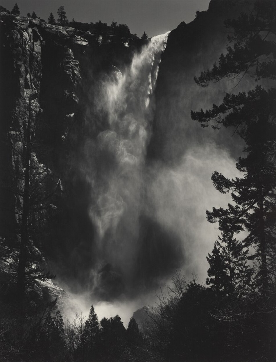 A misty waterfall drops vertiginously from a towering, sheer cliff face that nearly reaches the top edge of this vertical black and white photograph. Spray drifts in hazy clouds down the height and especially around the base of the falls, curling up into the trees that frame the view in front of us. Only a sliver of gray sky is shown above the cliff and falls.