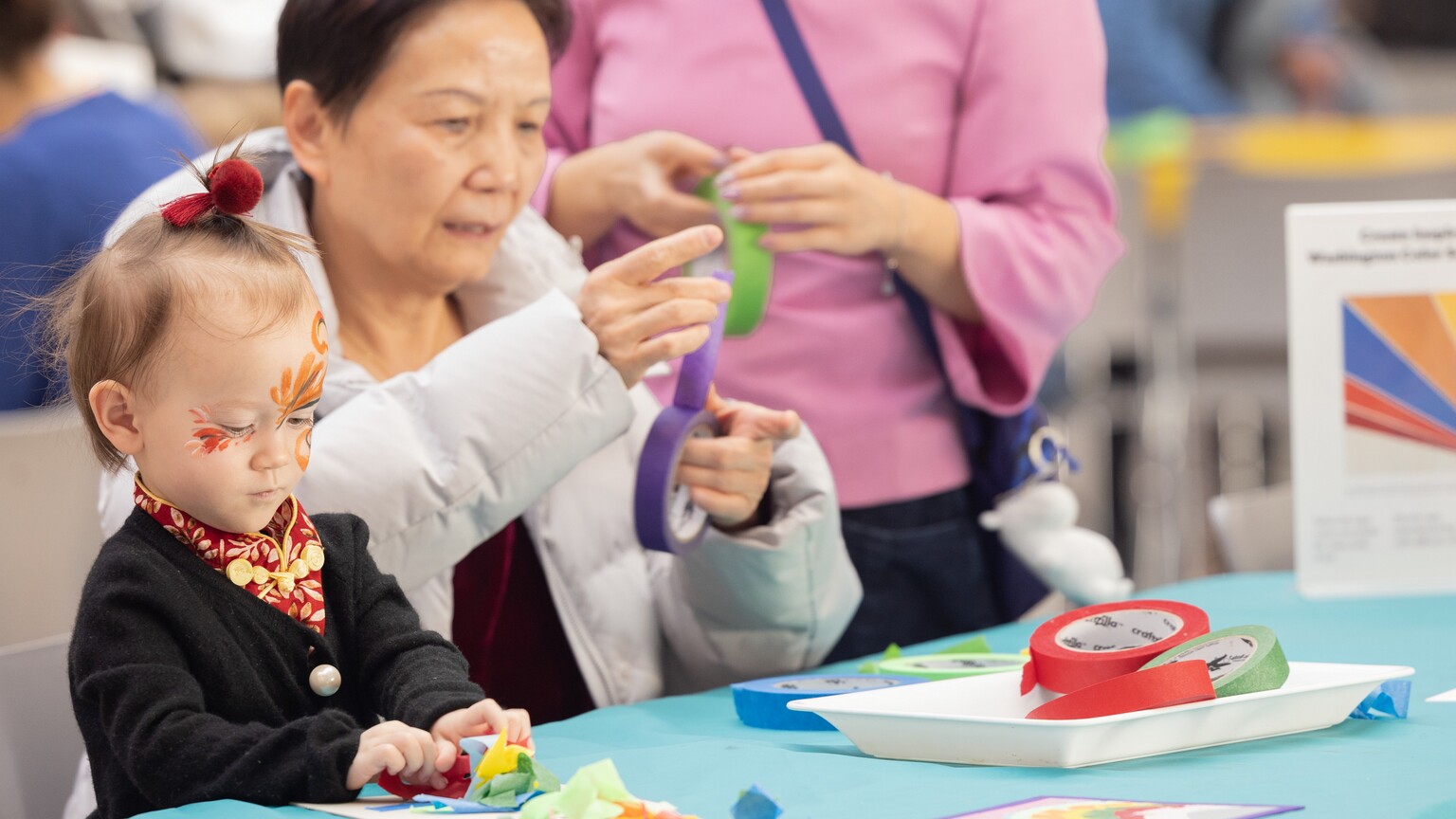 A little girl sitting at a table next to two women and all are working on arts and crafts projects.