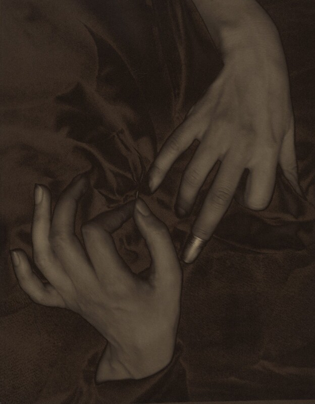 The backs of two splayed hands, palms down with one coming down from the top and one up from the bottom, nearly fill this vertical photograph and are shown lightly entwined in a field of dark fabric. The image is monochromatic like a black and white photograph but is printed in tones of golden and dark brown. The thumb of the left hand, coming up from the bottom, points upward so the fingers splay elegantly to our left. The forefinger and thumb touch to hold a needle stitching the dark fabric. The right hand comes down to our right and almost touches the left hand. The index finger is bent under at the first knuckle, and the ring and pinkie fingers gently curve downward into the fabric. The middle finger extends straight and is capped with a shiny thimble, which nearly touches the thumb of the opposite hand. The skin on the hands and wrists is smooth and the photograph is lightly blurred, creating the impression of a patina. The folds of the gathered fabric create a shadowy pinwheel effect.