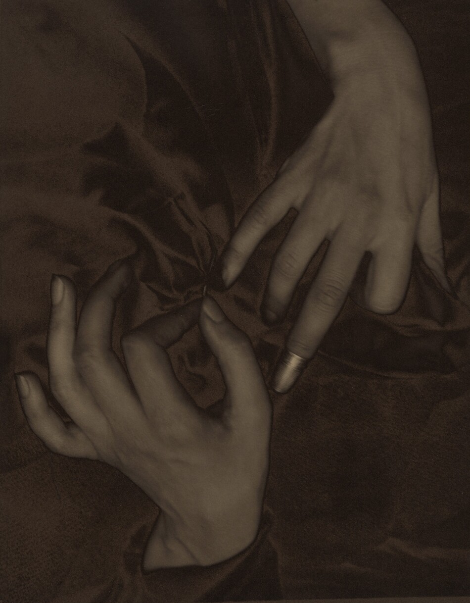 The backs of two splayed hands, palms down with one coming down from the top and one up from the bottom, nearly fill this vertical photograph and are shown lightly entwined in a field of dark fabric. The image is monochromatic like a black and white photograph but is printed in tones of golden and dark brown. The thumb of the left hand, coming up from the bottom, points upward so the fingers splay elegantly to our left. The forefinger and thumb touch to hold a needle stitching the dark fabric. The right hand comes down to our right and almost touches the left hand. The index finger is bent under at the first knuckle, and the ring and pinkie fingers gently curve downward into the fabric. The middle finger extends straight and is capped with a shiny thimble, which nearly touches the thumb of the opposite hand. The skin on the hands and wrists is smooth and the photograph is lightly blurred, creating the impression of a patina. The folds of the gathered fabric create a shadowy pinwheel effect.