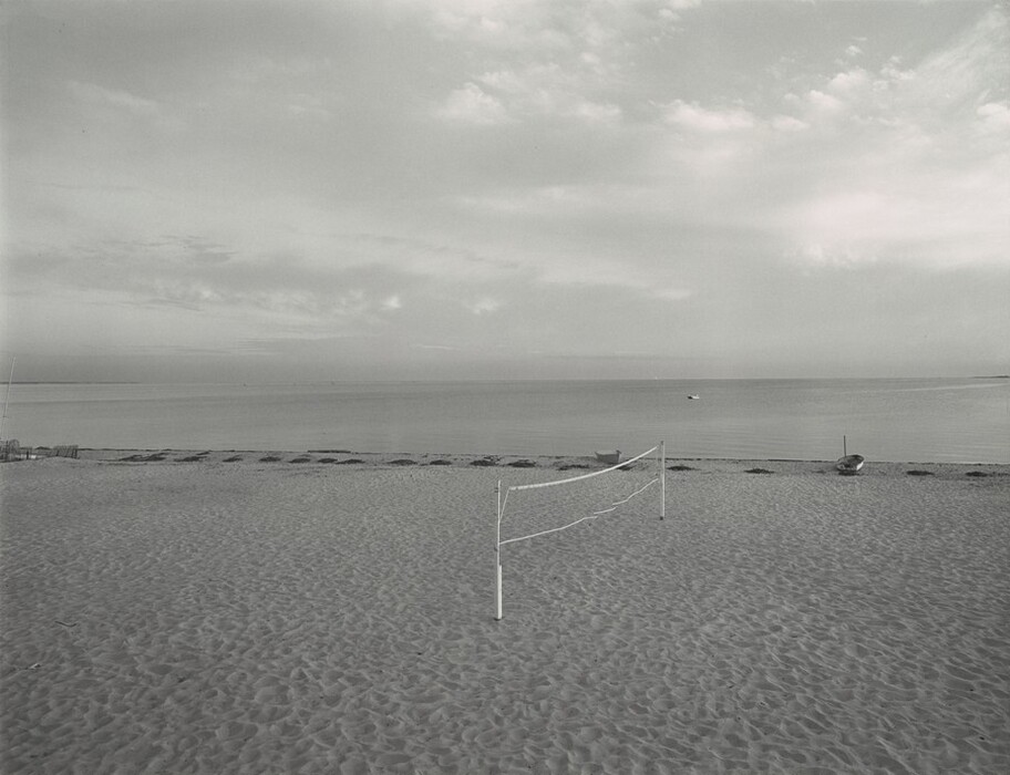 The image shows a beach with a calm sea. There is a wide sandy beach with footprints and ripples in the sand. A volleyball net is slightly off-center to the right. Seaweed runs parallel to the shoreline. In the distance, the water meets the horizon, with a small boat visible. The sky has a few wispy clouds. The image is in grayscale, with different shades of grey, creating a tranquil atmosphere.