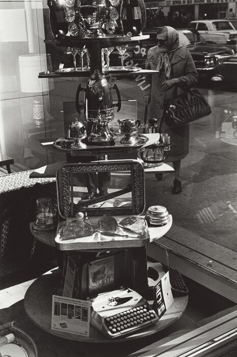The image shows a shop window with shelves filled with items, including a typewriter, mirrored objects, glassware, and a coffee pot. The image is in black and white, with a monochromatic color palette. In the foreground, a typewriter rests on the bottom shelf alongside magazines and metallic trays. The middle shelf displays glassware, and the top shelf showcases a large coffee pot and matching accessories. A street scene is reflected in the window, showing an elderly woman in a coat carrying a handbag and parked cars in the background.
