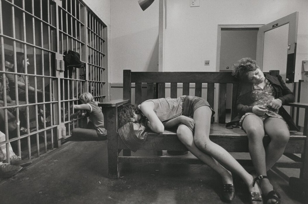 This is a photograph of a detention or holding cell area. The image shows a confined space with metal bars forming cells. Some individuals sit on the floor or lean against the bars. In the foreground, a wooden bench supports two subjects who appear to be asleep or resting, and one individual is sitting and leaning against the outside of the bars. The overall mood is one of waiting, tiredness, or distress, in the clinical institutional environment. The lighting is harsh, emphasizing the setting's bleakness.