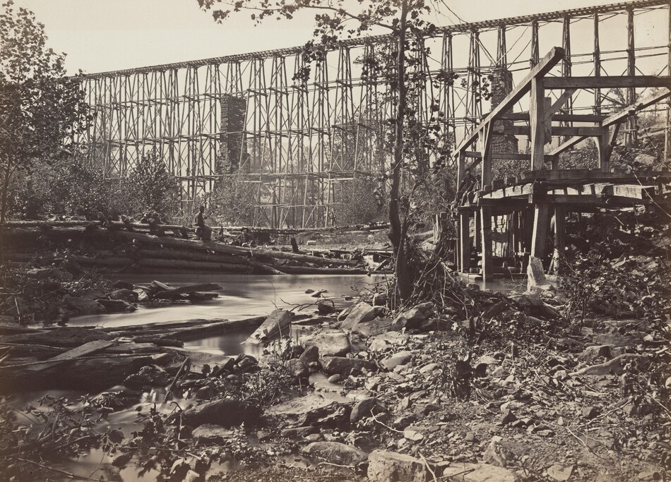 The image depicts a sepia-toned photograph of a large wooden trestle bridge extending across the scene. The sky appears clear with some brightness. A river flows near us, bordered by rocks and logs, with vegetation and trees visible in the background. To the right, a wooden structure runs parallel to the river, partially in shadow. A solitary figure sits on a log close to us beside the water. The overall tone of the photograph is earthy and monochromatic, lending a historic and timeless quality to the composition.