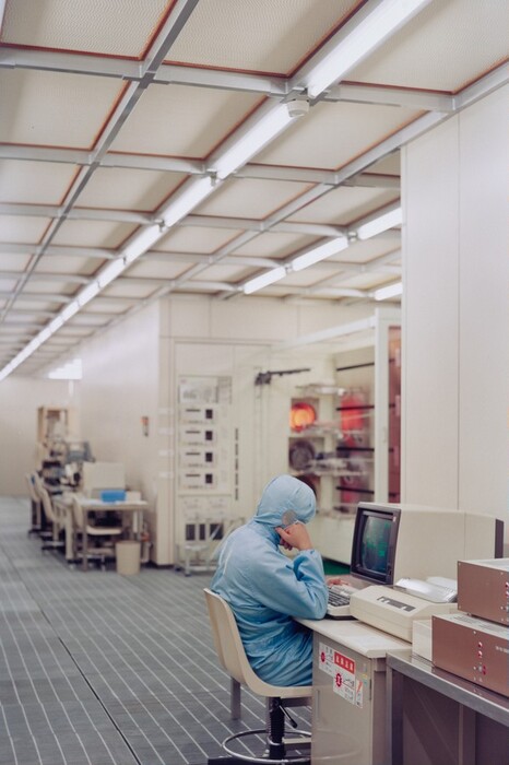 This is a photograph of a cleanroom environment. The image features an organized and sterile workspace typical in semiconductor or electronics manufacturing. The room is well-lit with fluorescent lights on the ceiling. A person in a full-body protective suit is seated at a desk with a vintage computer and electronic equipment. Around them are complex machinery and devices for a high-tech production process. The floor has anti-static mats and the walls are plain.