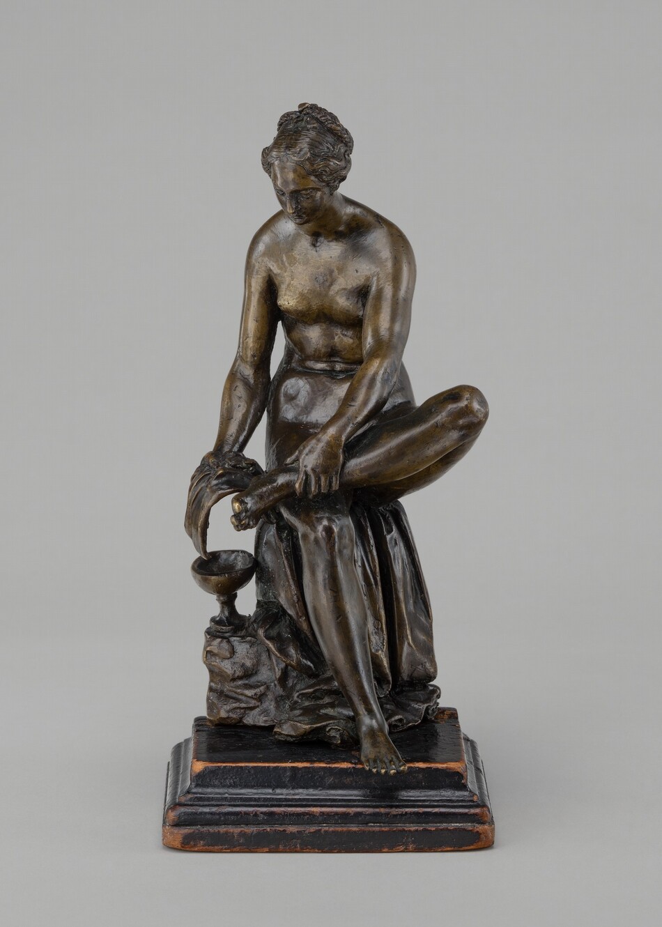 The sculpture depicts a seated nude female figure washing her feet. The figure is shown bending forward slightly, with one leg lifted and supported by her hands, directing water into a small basin or cup beneath her feet. The sculpture is made of bronze with intricate details, including flowing drapery covering part of her lower body. The figure's realistic posture, hair tied back, and focus on human anatomy are evident. The sculpture stands on a dark base, elevating the figure for visibility.