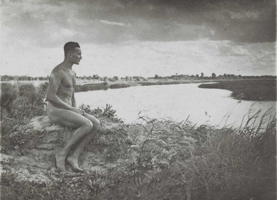 The image shows a person sitting on grass near a river. The person is seated with legs bent and facing the left side. The individual has short hair and is unclothed. The background includes water, trees, and vegetation under a cloudy sky.