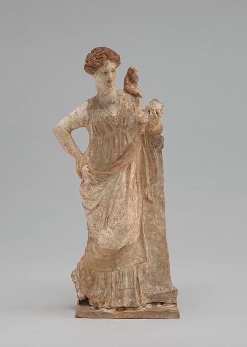 The sculpture depicts a woman standing with one hand on her hip and the other holding a small round object, while a bird or small animal perches on her shoulder. She seems to be leaning on a tall object on the right, which her bent elbow rests on. The woman is dressed in a draped gown that falls elegantly to her feet, with detailed folds in the fabric. Her skin is mostly white, with some patches of brown, and her reddish-brown hair is intricately styled on top of her head. Her outfit, the object she holds, and the creature on her shoulder are all blends of white, reddish-brown, and light brown pigments. The sculpture stands on a flat rectangular base.