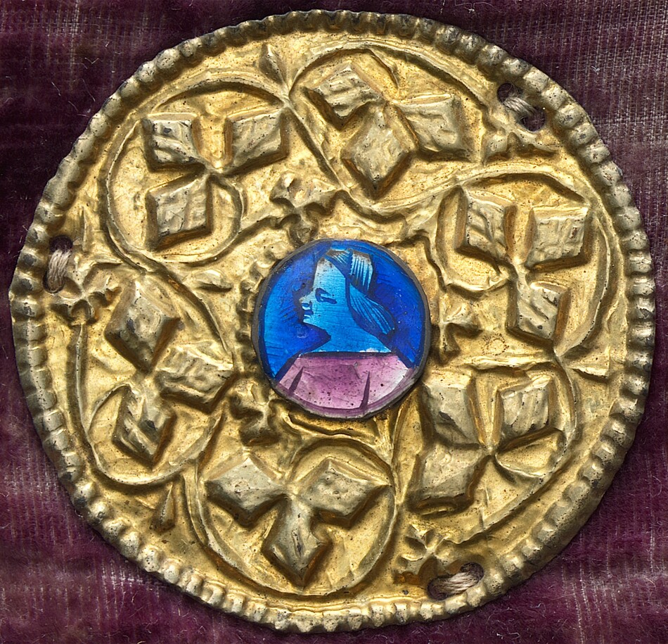 This is a photograph of an ornate medallion. The medallion is circular with a raised floral design engraved into a golden surface. At the center, there is an inset blue enamel piece featuring a stylized profile of a person. The background is a dark fabric.