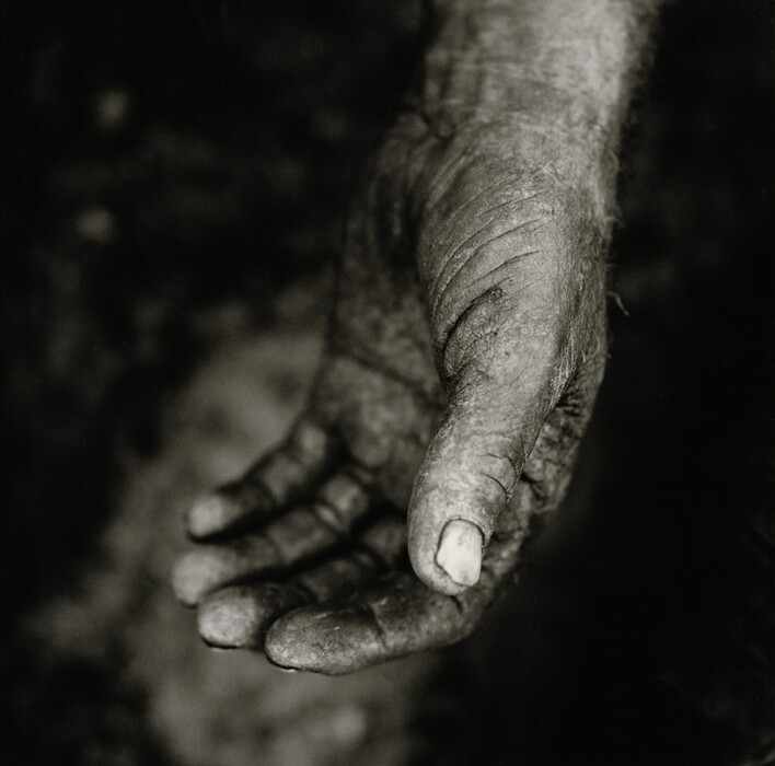 The image shows a close-up of a person's hand, extended slightly open towards the viewer. The hand displays signs of wear, with rough skin texture and dirt. The fingers and palm are prominently featured with the background out of focus and dark. The photograph is in black and white.
