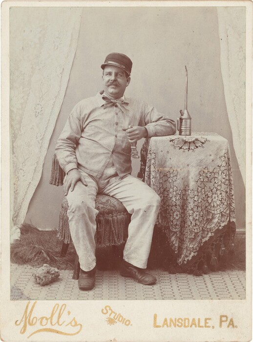 The image depicts a seated man wearing a checkered shirt, bow tie, white pants, dark shoes, and a cap. He is sitting on a fringed chair with one hand on his knee and the other on a table, holding a glass. An oil can is on the table and a small object is on the floor. The background consists of a plain wall with lace curtains.