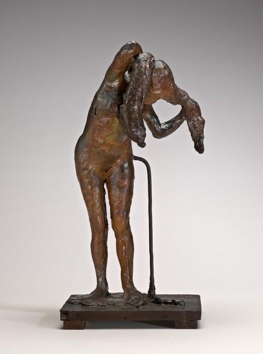 The sculpture depicts a figure of a person in a standing pose with their torso bent forward. The figure is holding a segment of long hair that hangs toward the floor. It appears to be a female form. The sculpture is composed of a bronze-like material with a dark patina, featuring rich earth tones such as deep browns and touches of lighter highlights. The surface of the sculpture is irregular and textured, capturing intricate details in the skin and hair. The figure stands on a rectangular base designed to look like a wooden platform, with a thin rod subtly attached to the figure along the side. Overall, the sculpture occupies space with realistic human proportions and an intimate engagement with natural human gestures.