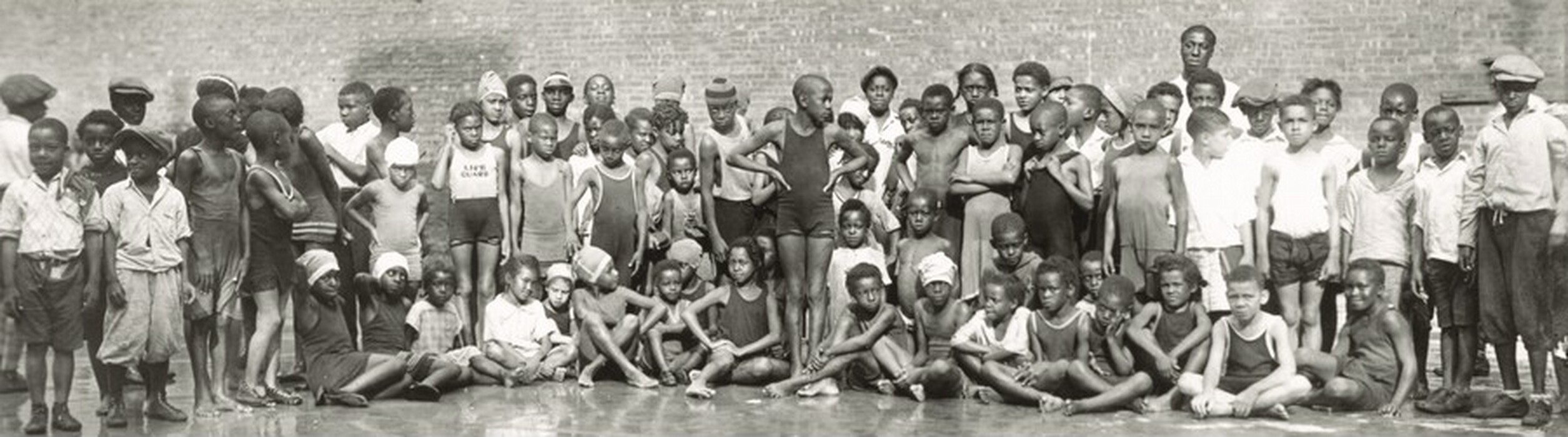 This long, horizontal, black-and-white photograph shows a group of Black children standing and sitting close together. Many look towards us, but some look at each other. The children vary in their poses, expressions, hairstyles, and clothing. Some wear caps on their heads. Some children are standing with arms by their sides, while others are sitting cross-legged. At the center of the image, one tall child stands up, their hands on their hips, looking to the right. On the right side, there appears to be an adult standing behind the long group of children. The background is a solid brick wall, and the gray floor is reflective.