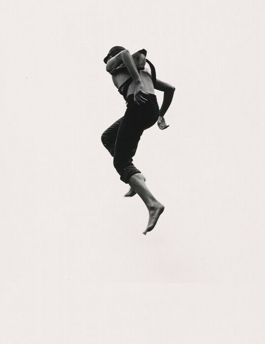 The image shows a person jumping in mid-air from a dynamic perspective. The person is in a side-on jumping position, with one arm bent and the other extended slightly back. Their face is not visible, and they are wearing dark pants rolled up at the ankles, a strappy top or suspenders, and appear to be barefoot. The background is plain and bright white.