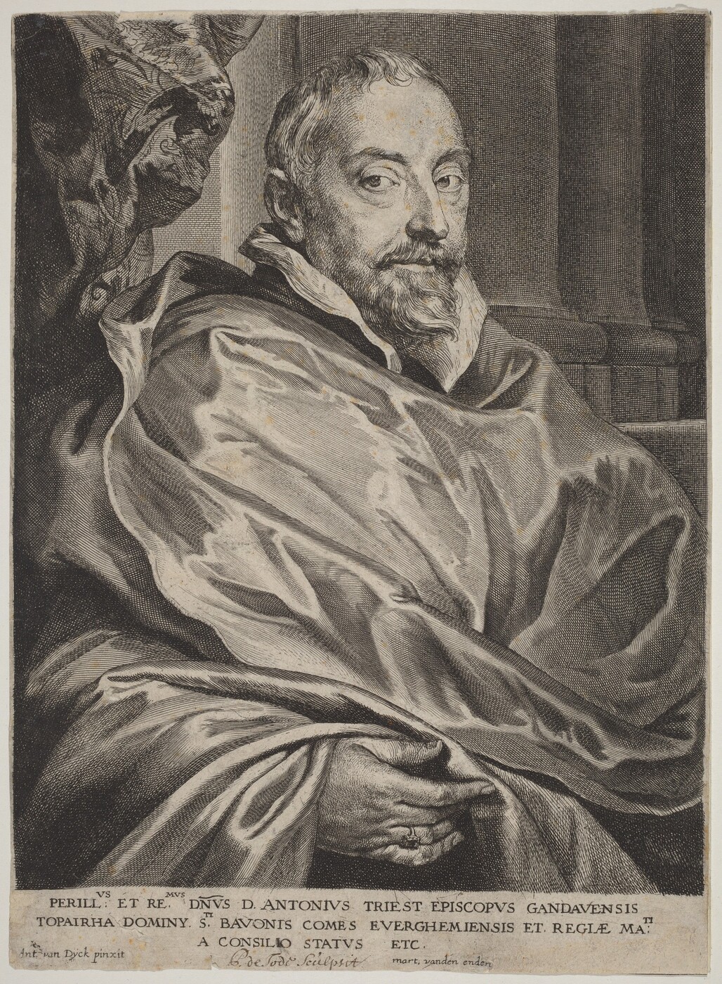 The image shows a seated man depicted from the waist up. He is positioned slightly turned to the side but faces forward. The man's facial features are detailed; he has a prominent, short beard and mustache, and his hair is short and neatly styled. He wears a flowing garment with voluminous folds, suggesting a robe or a cloak. The garment is draped around his shoulders. Stone architecture is visible in the background, indicating an interior space with columns. The man’s right hand is visible at the bottom of the image, and he is wearing a ring on his finger. A decorative cloth or tapestry is partially visible to the left of the image.