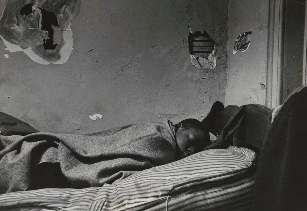 The image shows a young person lying on their side in bed, covered by a thick blanket that conceals most of their body, leaving only a portion of their face visible. Their eyes are closed, indicating they are asleep. The person has short hair, and their head rests on a striped pillow. There are no visible jewelry or objects in their hands. The background features a wall with visible damage, including peeling paint and holes that reveal lath strips beneath. The bed is positioned near a window, which is partially visible to the right side of the image.