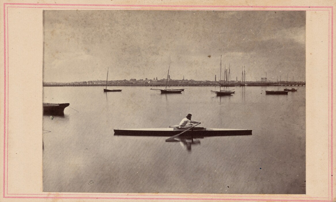 The image shows a person rowing a long boat on a calm body of water, with sailboats scattered in the background. A cityscape is visible on the elevated horizon, reflecting in the water. The sky is overcast, creating a muted light over the scene. The color palette is monochromatic with sepia tones.