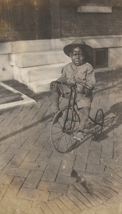 The image shows a young child sitting on a tricycle. The child is seated with hands on the handlebars, slightly turned to the side. The child has a neutral or slight smile, short hair covered by a hat, and wears a matching jacket, shorts, socks, and shoes. The ground is made of rectangular stone pavers, and in the background, there is a structure with steps and a brick wall. The setting appears to be outdoors in front of a building.