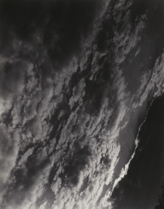 The image displays a view of the sky with dynamic cloud formations, shot from a slightly tilted perspective. The horizon is not visible, solely focusing on the movement of the clouds above. The stark contrasts between light and shadow capture the texture and depth of the clouds. The color palette is monochromatic with primarily black and white shades, emphasizing the clouds' boldness. Darker areas show the density and shape of the clouds, while lighter areas suggest potential sunlight. In the distance, the clouds merge and swirl, creating a dance of light and form against the vast sky.