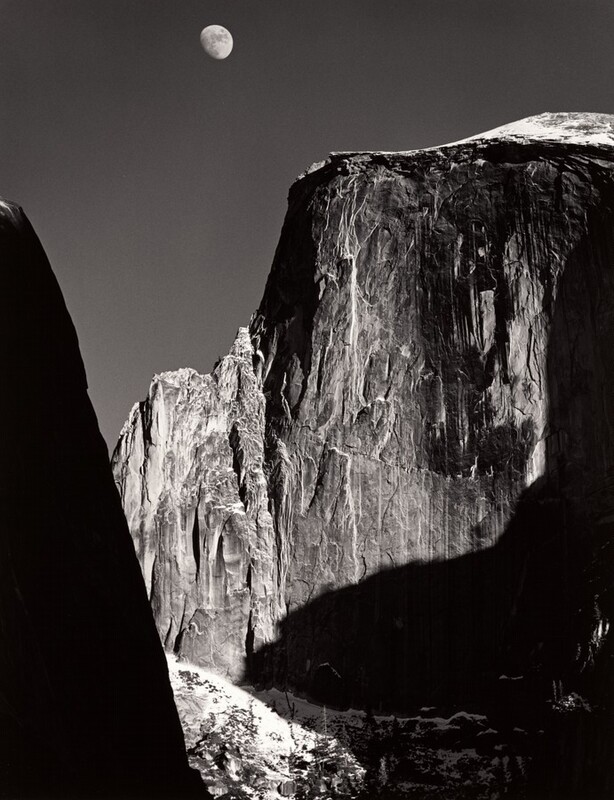 From a low vantage point, we look up at the rugged face of a tall, almost sheer rock formation with the moon above in this vertical black and white photograph. Filling most of the composition, the rock formation is slightly rounded along the top but dips down to a jagged ridge to our left. Strong light from the upper right creates deep shadows across its iron-gray surface, which is textured with long vertical streaks of white and lighter gray. An almost-full moon hovers in the otherwise clear, charcoal-gray sky above it, near the top of the image to our left of center. Formations outside our view cast deep shadows along the right edge of the photograph. A smattering of white across from us, low in the image, suggest snow on the rock there. A dark mass close to us is an ink-black silhouette rising from the lower left corner and angling off the left edge, near the upper left corner.