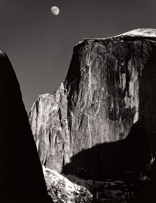 From a low vantage point, we look up at the rugged face of a tall, almost sheer rock formation with the moon above in this vertical black and white photograph. Filling most of the composition, the rock formation is slightly rounded along the top but dips down to a jagged ridge to our left. Strong light from the upper right creates deep shadows across its iron-gray surface, which is textured with long vertical streaks of white and lighter gray. An almost-full moon hovers in the otherwise clear, charcoal-gray sky above it, near the top of the image to our left of center. Formations outside our view cast deep shadows along the right edge of the photograph. A smattering of white across from us, low in the image, suggest snow on the rock there. A dark mass close to us is an ink-black silhouette rising from the lower left corner and angling off the left edge, near the upper left corner.