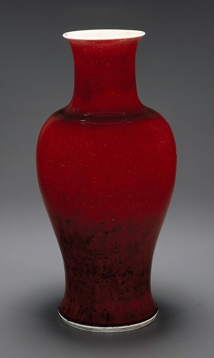 This is a vase with a vibrant red glaze. The vase has a narrow neck, a slightly wider body, and a slightly flared rim. Its surface appears smooth. The bottom of the vase has a subtle gradient, with the red fading into black. The rim of the base appears to be bright white.