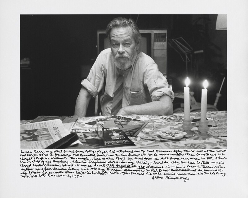 A pale-skinned, bearded man sits on the far side of a table strewn with magazines and newspapers in this horizontal, black and white photograph. The man’s shoulders are angled slightly to our left, and he looks steadily out at us with hooded eyes. Wrinkles line his forehead and cheeks between his nose and mouth. His straight hair is brushed up and back and he has a full beard. His left forearm, to our right, lies along the edge of the table, and he leans onto that elbow. The other hand rests on his knee. His long shirt sleeves are rolled up over the elbows, and he wears a diagonally striped tie. Something weighs down the breast pocket of his rumpled shirt. Two lit taper candles are in short, bulb-shaped candlesticks to the right next to an ashtray holding cigarette butts. The foil surface of a cigarette box gleams on the table behind the candles. A pair of glasses sits on two magazines in front of the man. A box television is directly behind the man, and other objects in the background are swallowed in shadow. A long, handwritten inscription along the bottom reads, “Lucien Carr, my oldest friend from college days, had introduced me to Jack Kerouac after they’d met at the West End bar on 113th St. & Broadway, and connected Jack + me to his fellow St. Louis uppermiddle-class (aristocrat we thought) confrère William S. Burroughs, late winter 1944. We lived down the hall from each other on 7th floor Union Theological Seminary, Columbia freshman dorm during W.W.II, I heard haunting Brahms sextet music through his door, knocked, we met. Kerouac heard _Old Angel Midnight_ eloquence in Lucien’s laconic “Celtic” intonations. Here four decades later, wise Old Dog Bureau Manager United Press International he was visiting former house-mate Alene Lee’s Soho loft. More eyes perused his wire-service prose than all Jack’s & my texts, I’ll bet. December 2, 1986. Allen Ginsberg.”