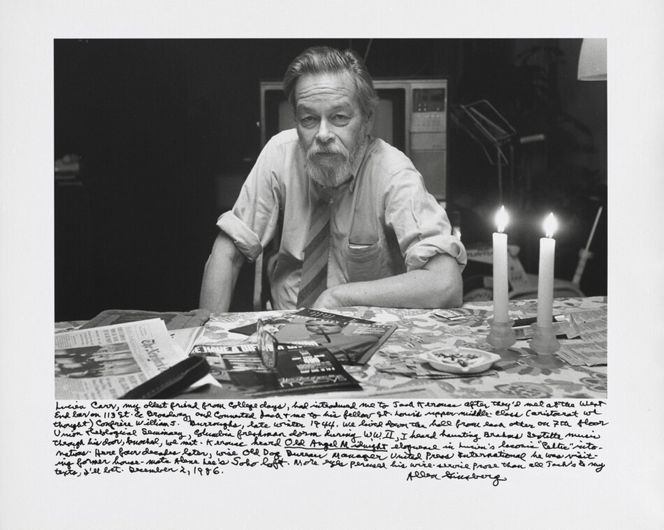 A pale-skinned, bearded man sits on the far side of a table strewn with magazines and newspapers in this horizontal, black and white photograph. The man’s shoulders are angled slightly to our left, and he looks steadily out at us with hooded eyes. Wrinkles line his forehead and cheeks between his nose and mouth. His straight hair is brushed up and back and he has a full beard. His left forearm, to our right, lies along the edge of the table, and he leans onto that elbow. The other hand rests on his knee. His long shirt sleeves are rolled up over the elbows, and he wears a diagonally striped tie. Something weighs down the breast pocket of his rumpled shirt. Two lit taper candles are in short, bulb-shaped candlesticks to the right next to an ashtray holding cigarette butts. The foil surface of a cigarette box gleams on the table behind the candles. A pair of glasses sits on two magazines in front of the man. A box television is directly behind the man, and other objects in the background are swallowed in shadow. A long, handwritten inscription along the bottom reads, “Lucien Carr, my oldest friend from college days, had introduced me to Jack Kerouac after they’d met at the West End bar on 113th St. & Broadway, and connected Jack + me to his fellow St. Louis uppermiddle-class (aristocrat we thought) confrère William S. Burroughs, late winter 1944. We lived down the hall from each other on 7th floor Union Theological Seminary, Columbia freshman dorm during W.W.II, I heard haunting Brahms sextet music through his door, knocked, we met. Kerouac heard _Old Angel Midnight_ eloquence in Lucien’s laconic “Celtic” intonations. Here four decades later, wise Old Dog Bureau Manager United Press International he was visiting former house-mate Alene Lee’s Soho loft. More eyes perused his wire-service prose than all Jack’s & my texts, I’ll bet. December 2, 1986. Allen Ginsberg.”