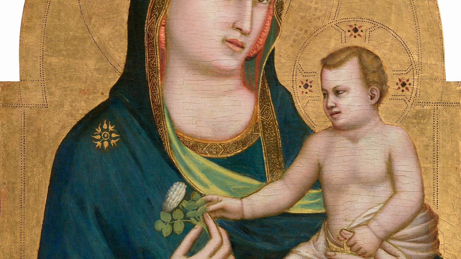 A woman with pale skin holds a baby in the crook of her left arm, on our right, against a gold background in this vertical, arched wooden panel. The woman is shown from the waist up with her body angled slightly to our right. She wears a dark blue mantle that drapes over her head and shoulders, and across her body. The garment has a gold border and celery-green lining where it turns over at her wrists and chest. There is a gold starburst-like symbol on her right shoulder, our left, and she holds a stylized rose with her right hand. The baby is nude except for a translucent white cloth, also edged with a gold border, wrapped around his waist and legs. His face and body look more like a small man than a baby, but there are baby-like rolls at his wrists. He has blond wavy hair. He grips the woman's forefinger on the hand that holds his body, and he reaches for the rose she holds with his other hand. The gold background is punched to create halos around their heads and decorative bands along the inner edge of the curving, arched top.