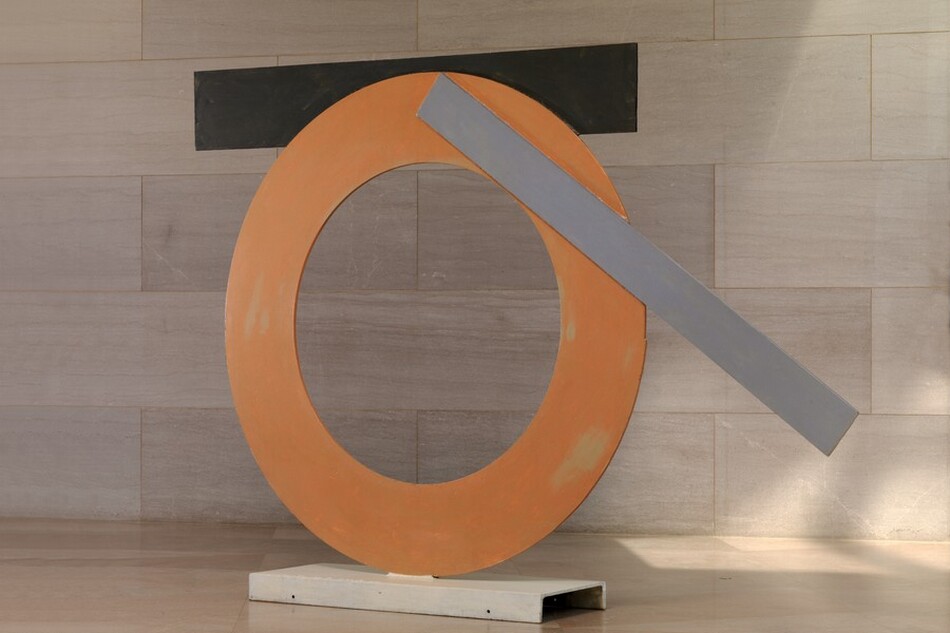 Made with sheets of painted steel, an orange ring stands upright on a narrow white base in this freestanding sculpture. A black strip and a gray strip are affixed to the edges of the ring. The black strip is horizontal across the top of the circle, attached to the back side of the ring, and the narrower but longer gray strip is angled across the top right quarter of the circle. The sculpture is photographed near a wall clad with pale pinkish-gray marble and a similarly colored floor.