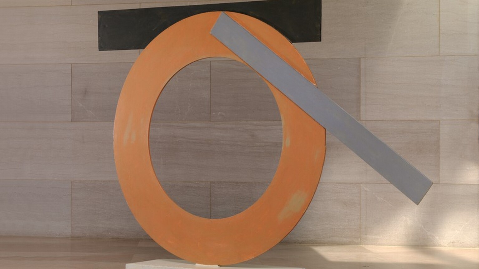 Made with sheets of painted steel, an orange ring stands upright on a narrow white base in this freestanding sculpture. A black strip and a gray strip are affixed to the edges of the ring. The black strip is horizontal across the top of the circle, attached to the back side of the ring, and the narrower but longer gray strip is angled across the top right quarter of the circle. The sculpture is photographed near a wall clad with pale pinkish-gray marble and a similarly colored floor.