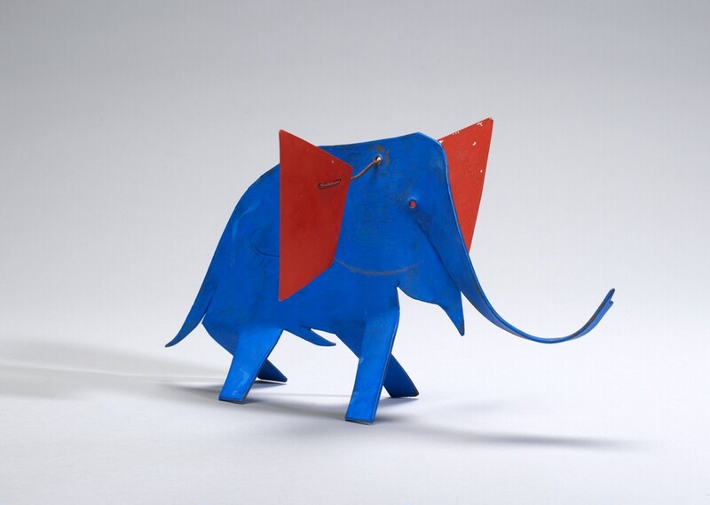 The form of an elephant is made with metal sheets colored lapis-blue and scarlet red in this freestanding sculpture. The four legs are flat strips that have been bent in alternating directions to support the sculpture in a wide-legged stance. The trunk curls up and the trapezoidal ears are connected with a metal wire threaded through a hole in the animal’s head so they balance on either side. The sculpture casts faint shadows on the white surface on which it is photographed.
