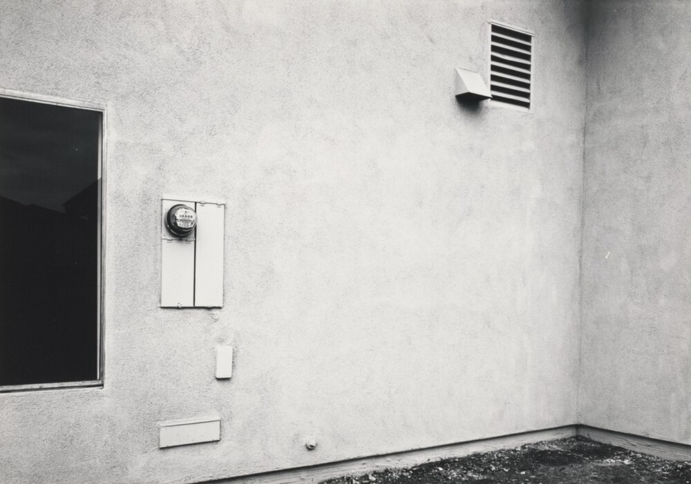 This is a photograph of a plain exterior wall with a few structural elements. The photo shows a wall with a flat, textured surface in a grayish tone. On the left side, there is a dark, reflective window. Next to the window is a utility meter enclosed in a metal casing. Above the meter, there is a slanted vent cover. The wall meets the bare ground in the image.