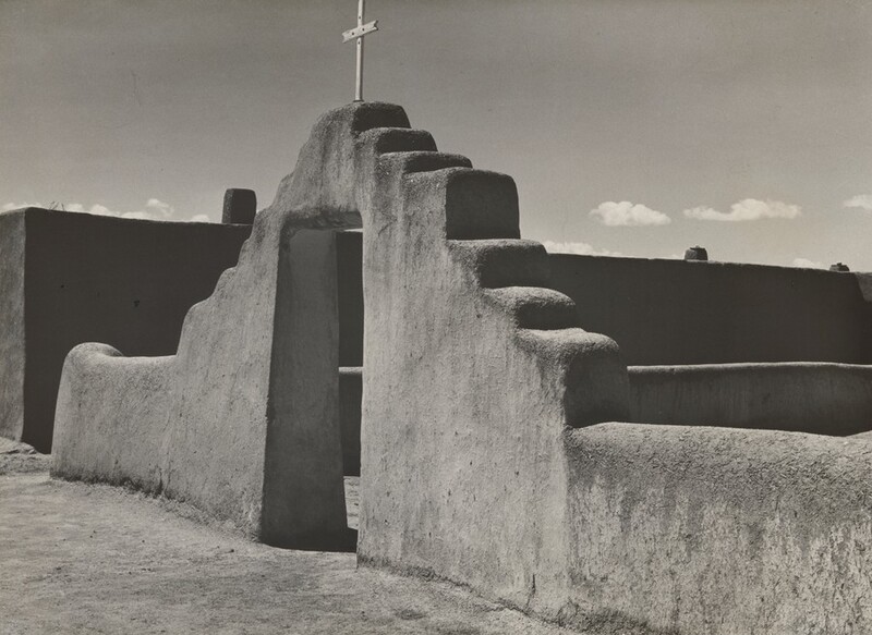 This grayscale photograph shows an adobe-style doorway with steps leading up to a cross at the top, set against a sky with a few small clouds. There are low walls on either side of the doorway. The adobe texture appears rough and coarse. Behind the central doorway is another wall or building that is in shadow.
