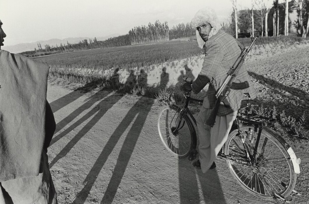 This is a photograph of a dirt path with a bicycle and fields in the background. The image captures a bicycle on a dirt path, surrounded by fields and a line of trees that recede into the distance. Shadows cast by the sun fall long across the ground stretching toward the fields. The ground appears dry with sparse vegetation at the edges. Layers of distant mountains can be seen in the horizon, creating depth in the composition.