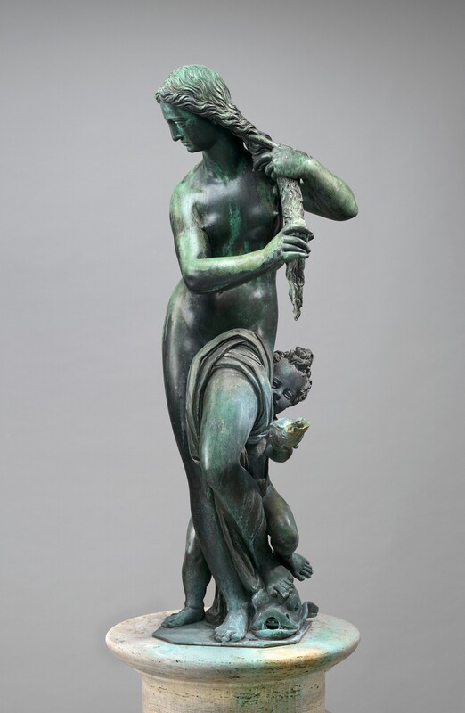 A nearly nude woman wrings out her long hair as a young boy peeks around one hip in this free-standing bronze fountain. In this photograph, the woman's hips and shoulders are squared toward us. Her face turns to our left, and she looks down in that direction so we see her in profile. She gathers her curly hair in a long tress and wrings it with both hands. Her right leg, to our left, is straight, and her other knee is bent so that foot rests on a stylized fish. Cloth drapes over the top of the bent thigh to cover her groin and falls between her legs to her ankles. The fish faces us with its mouth open and two canine-like teeth flanking a hole where water would have spouted. The child gazes around her left hip, to our right, so his eyes, snub nose, and one round cheek angle into view. He has a pile of short curls atop his head, and he holds out a conch shell with one hand. His feet straddle the woman’s in a wider stance so his left foot, to our right, rests with hers over the fish. The patina of the sculpture's polished bronze surface has turned minty green in some areas, especially on the back of the woman’s head, her arms, hands, along her left side, over the bent thigh, and the shell the boy holds. Other areas are darker gray. The pair stand on a piece of flat metal, which is on an off-white, stone pedestal against a light gray background.