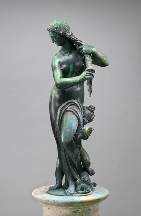 A nearly nude woman wrings out her long hair as a young boy peeks around one hip in this free-standing bronze fountain. In this photograph, the woman's hips and shoulders are squared toward us. Her face turns to our left, and she looks down in that direction so we see her in profile. She gathers her curly hair in a long tress and wrings it with both hands. Her right leg, to our left, is straight, and her other knee is bent so that foot rests on a stylized fish. Cloth drapes over the top of the bent thigh to cover her groin and falls between her legs to her ankles. The fish faces us with its mouth open and two canine-like teeth flanking a hole where water would have spouted. The child gazes around her left hip, to our right, so his eyes, snub nose, and one round cheek angle into view. He has a pile of short curls atop his head, and he holds out a conch shell with one hand. His feet straddle the woman’s in a wider stance so his left foot, to our right, rests with hers over the fish. The patina of the sculpture's polished bronze surface has turned minty green in some areas, especially on the back of the woman’s head, her arms, hands, along her left side, over the bent thigh, and the shell the boy holds. Other areas are darker gray. The pair stand on a piece of flat metal, which is on an off-white, stone pedestal against a light gray background.