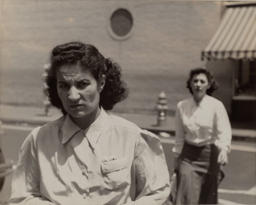 The image depicts two women outdoors. One woman is in the foreground, walking towards the camera. She has curly dark hair styled back, a serious expression, and is wearing a white blouse with a pointed collar. The other woman is in the background, also walking. The background includes an urban cityscape with a building, a circular window, a striped awning, a sidewalk, and a hydrant.