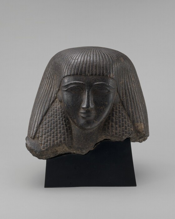 The sculpture depicts the head of a person in ancient Egyptian style. The person has a large forehead, almond-shaped eyes, and a small smile. The hair is shown as a thick wig with straight bangs and long strands falling to the shoulders. Carved from dark stone, the sculpture has a smooth surface with a subtle sheen. There are signs of wear around the edges, suggesting age and possibly that it is part of a larger artwork. The hair is detailed with horizontal lines and a textured pattern towards the ends, adding to the lifelike but formalized appearance typical of ancient Egyptian art.