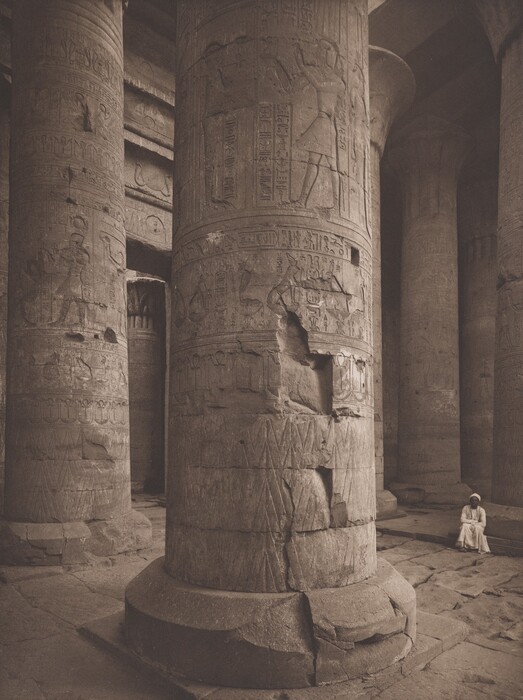 This is a photograph showing intricately carved stone columns within an ancient structure. The image displays a series of massive stone columns adorned with hieroglyphics and detailed carvings of figures and symbolic elements. The columns are located in an open, spacious area of a historical site. The carvings depict a narrative, showcasing the antiquity and cultural significance of the place. The floor consists of large stone slabs, and light filtering into the space creates a subdued, rustic atmosphere, highlighting the texture and age of the stone. The photograph captures the grandeur and historical depth of ancient architecture.