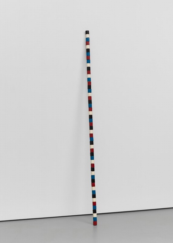 Fifty-two straight-sided wood beads in white, black, blue, and red are stacked, presumably around a dowel or wire, so the resulting staff-like column stands on a gray floor and leans against a white wall. The beads do not form a regular pattern but each color is represented in each grouping of four beads.