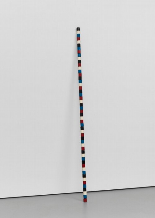 Fifty-two straight-sided wood beads in white, black, blue, and red are stacked, presumably around a dowel or wire, so the resulting staff-like column stands on a gray floor and leans against a white wall. The beads do not form a regular pattern but each color is represented in each grouping of four beads.