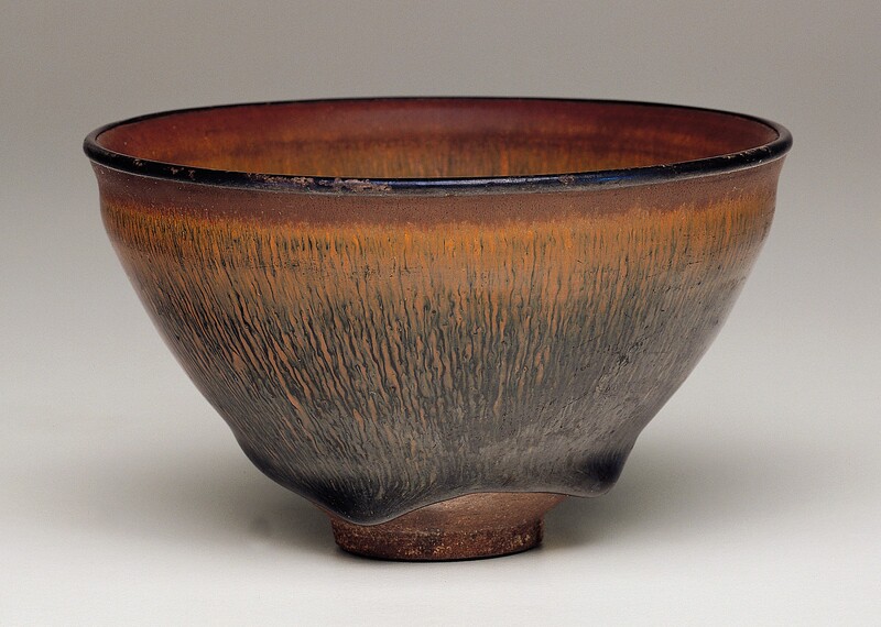 This round ceramic bowl has a distinctive brown glaze. The lip is black and the rim immediately under it is bronze brown. From there, copper-orange rivulets trickle down the sides of the bowl and intermingle with black veins below. The bottom of the bowl has an uneven, wavy base. The bowl is photographed against a background that lightens from gray along the top to white along the bottom.