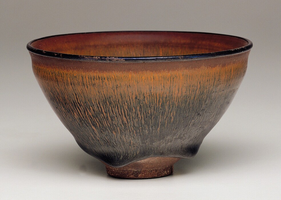This round ceramic bowl has a distinctive brown glaze. The lip is black and the rim immediately under it is bronze brown. From there, copper-orange rivulets trickle down the sides of the bowl and intermingle with black veins below. The bottom of the bowl has an uneven, wavy base. The bowl is photographed against a background that lightens from gray along the top to white along the bottom.