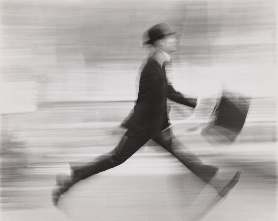 This is a photograph of a person in mid-stride, holding a briefcase. The image shows motion blur effect. The figure is dressed in formal attire, including a hat. The background is blurred, suggesting an urban setting. The photo is in monochrome.