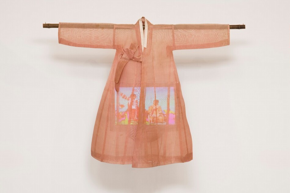 A lightweight, sheer, coral-peach robe hangs from a wooden dowel over a color television screen in this sculptural piece, which hangs against a white wall. The robe has long sleeves extending horizontally along the dowel, which extends a bit past the cuffs. The robe has a narrow, cream-white collar and ties to one side over the chest. It flares slightly below the arms, creating a bell shape. The rectangular video screen that hangs within is partially obscured, but looks like at least two people in a brightly colored setting.