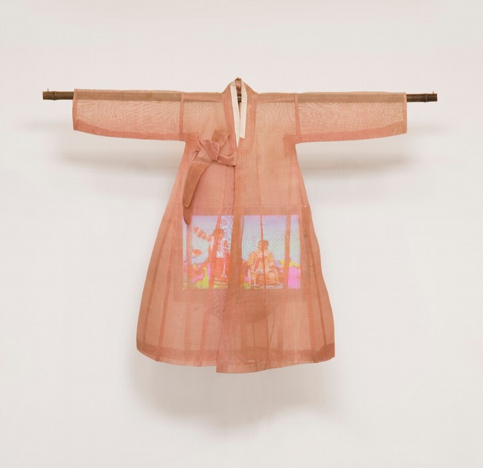 A lightweight, sheer, coral-peach robe hangs from a wooden dowel over a color television screen in this sculptural piece, which hangs against a white wall. The robe has long sleeves extending horizontally along the dowel, which extends a bit past the cuffs. The robe has a narrow, cream-white collar and ties to one side over the chest. It flares slightly below the arms, creating a bell shape. The rectangular video screen that hangs within is partially obscured, but looks like at least two people in a brightly colored setting.