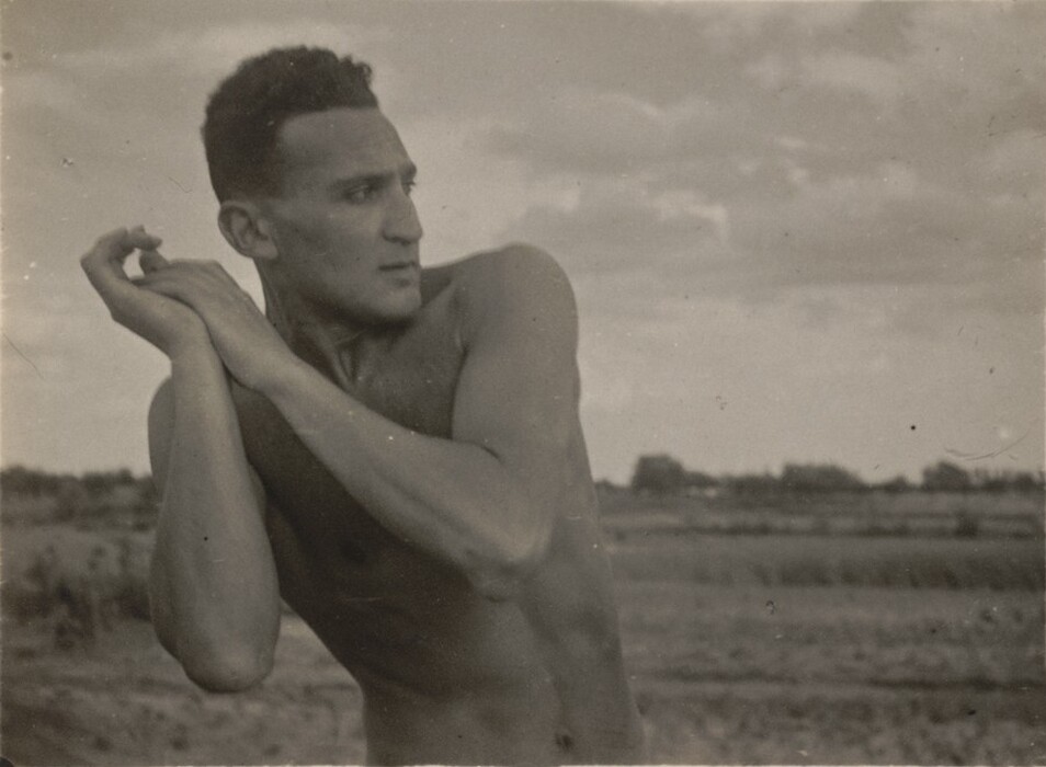 The image shows an upper body portrait of a man, featuring his torso, arms, and head. He is posed with his arms raised, hands clasped near his face, and his head turned slightly to the side, looking away from the camera. The man's facial features include a strong jawline, a straight nose, and a focused expression. He has short, curly hair. He appears to be shirtless, and there are no visible clothing items. The background is an open outdoor landscape with fields and scattered trees under a cloudy sky. The photo is in black and white.