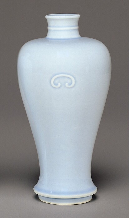 The sculpture depicts a vase made of porcelain with an elongated form that widens gently into a rounded midsection before tapering towards the base. The vase has a light blue glaze covering its surface and a decorative swirl motif embossed on the upper front.